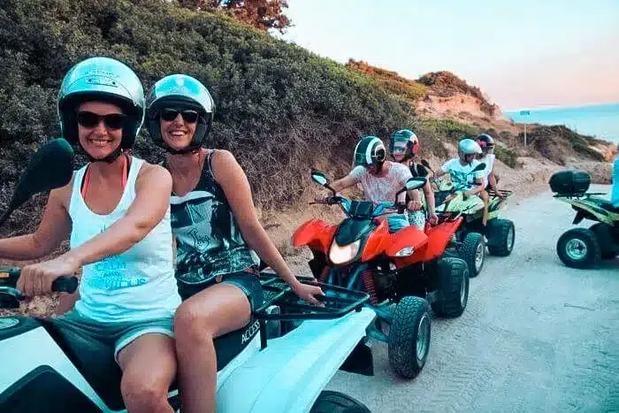 Zwei Menschen mit Helmen lächeln, während sie auf Geländefahrzeugen auf einem staubigen Pfad sitzen, im Hintergrund eine malerische Aussicht auf die Küste. Weiter entlang des Pfads sind weitere Fahrer zu sehen.