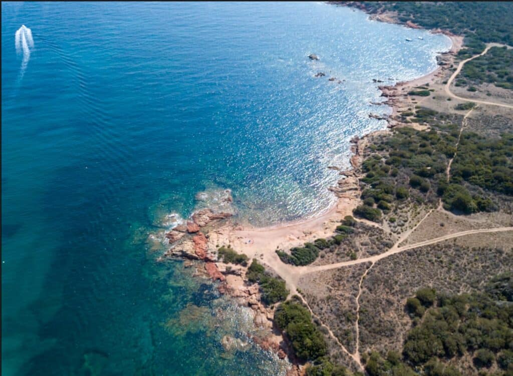 Luftaufnahme einer zerklüfteten Küste mit glitzerndem türkisfarbenem Wasser, das auf ein felsiges Ufer trifft, neben einem von Bäumen gesäumten Pfad und einem Schnellboot, das während eines Segeltörns eine weiße Kielwelle im Meer erzeugt.