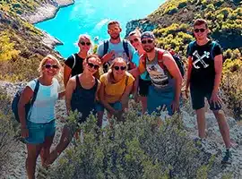 Eine Gruppe von sieben Freunden lächelt an einem sonnigen Tag auf einem Hügel und blickt auf einen blauen Fluss, der sich während ihres Segeltörns durch eine felsige Landschaft schlängelt.