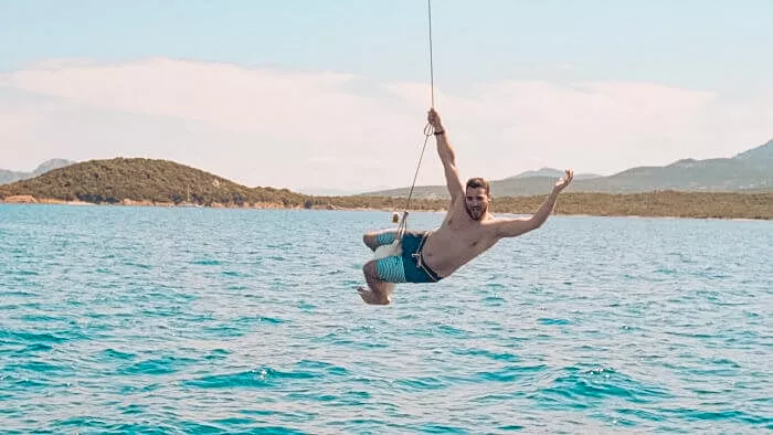 Ein Mann schwingt während eines sonnigen Segelurlaubs fröhlich auf einem Seil über einem klaren, blauen Meer mit malerischen Hügeln im Hintergrund.