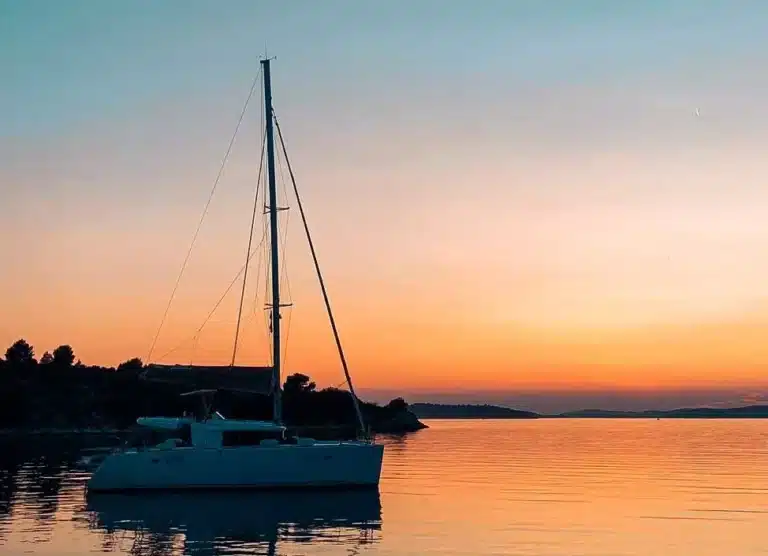 Eine Segelyacht, die auf ruhigen Gewässern vor einem Sonnenuntergangshimmel mit orangefarbenen und rosa Farbtönen verankert ist, die sich sanft im Wasser spiegeln.