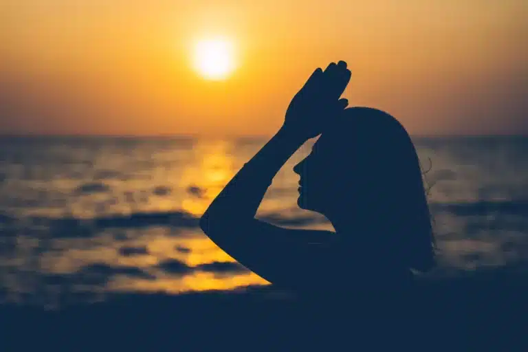 Silhouette einer Frau am Meer bei Sonnenuntergang, ihre Hand ruht anmutig auf ihrer Stirn, im Hintergrund geht die Sonne über dem Wasser unter, während sie ein Segelboot in der Ferne beobachtet.