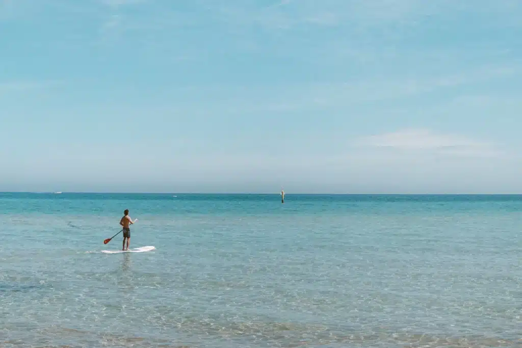 Eine Person fährt mit dem Paddleboard auf einem ruhigen, klaren Meer unter einem weiten blauen Himmel, mit einem dünn besiedelten Horizont und einer in der Ferne sichtbaren Segelyacht.