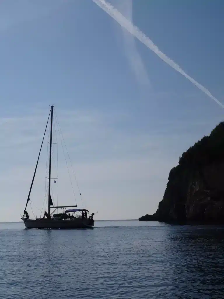 Eine Segelyacht schwimmt unter klarem Himmel in der Nähe einer felsigen Küste, über ihr ist ein Kondensstreifen eines Flugzeugs sichtbar.