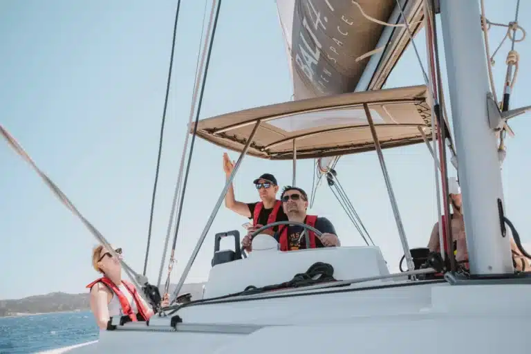 Drei Personen genießen einen sonnigen Tag auf einer Segelyacht. Ein Mann steuert, während eine Frau und ein anderer Mann zuschauen. Alle tragen Schwimmwesten und Sonnenbrillen.