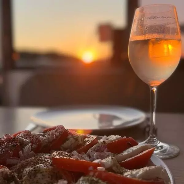 Eine Nahaufnahme einer köstlichen Mahlzeit mit Tomaten und Käse auf einem Teller, neben einem Glas Wein, auf einer Segelyacht mit einem wunderschönen Sonnenuntergang im Hintergrund.