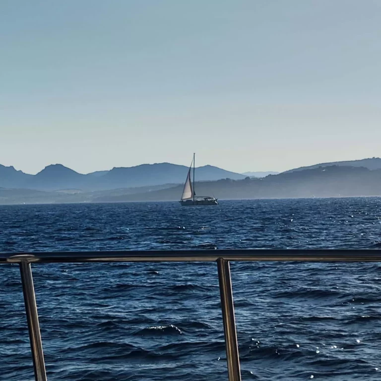 Eine Segelyacht kreuzt auf dem offenen Meer, sichtbar durch die Metallreling des Bootes, mit Bergen in der Ferne unter einem klaren blauen Himmel.