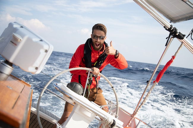 Skipper Max von Sailwithus mit roter Jacke und Sonnebrielle an Bord der modernen Segelyacht mit guter Laune auf rauher See