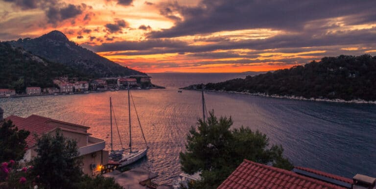Ein Panoramablick auf ein Küstendorf bei Sonnenuntergang, mit den Silhouetten der festgemachten Segelboote bereit für eine Segelreise vor einem lebendigen Himmel mit Wolken, umgeben von üppigen Hügeln und Ruhe