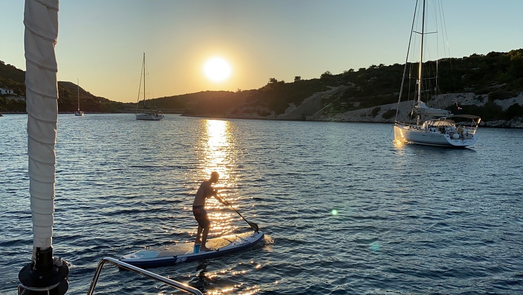 Eine Person fährt bei Sonnenuntergang auf einem ruhigen Meer Paddleboard, in der Nähe ankern Segelboote und im Hintergrund sind Hügel unter einem goldenen Himmel zu sehen.