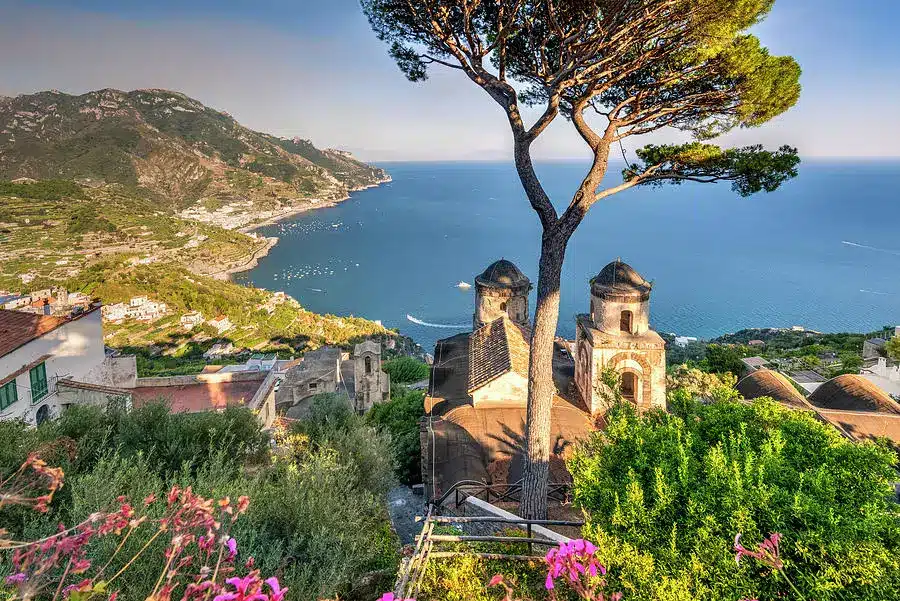 Ein atemberaubender Blick auf eine Küstenlandschaft mit einer historischen Kirche mit zwei Glockentürmen, umgeben von üppigem Grün und einem großen Baum, mit einem klaren blauen Meer und einer zerklüfteten Küste im Hintergrund, perfekt für