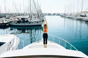 Eine Frau steht auf dem Bug einer Segelyacht, streckt ihre Arme zum Himmel und blickt auf einen ruhigen Yachthafen voller angedockter Segelboote unter einem klaren blauen Himmel.