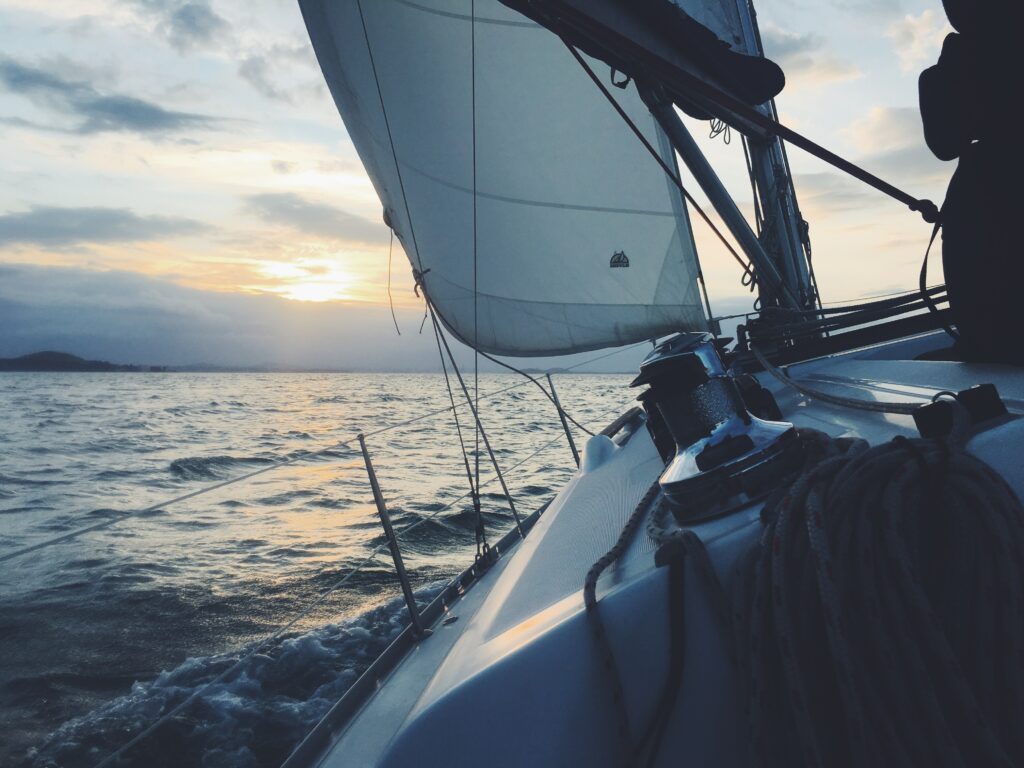 Ein Katamaran fährt bei Sonnenuntergang mit gesetzten Segeln auf ruhiger See vor der Kulisse vereinzelter Wolken und einer untergehenden Sonne am Horizont.