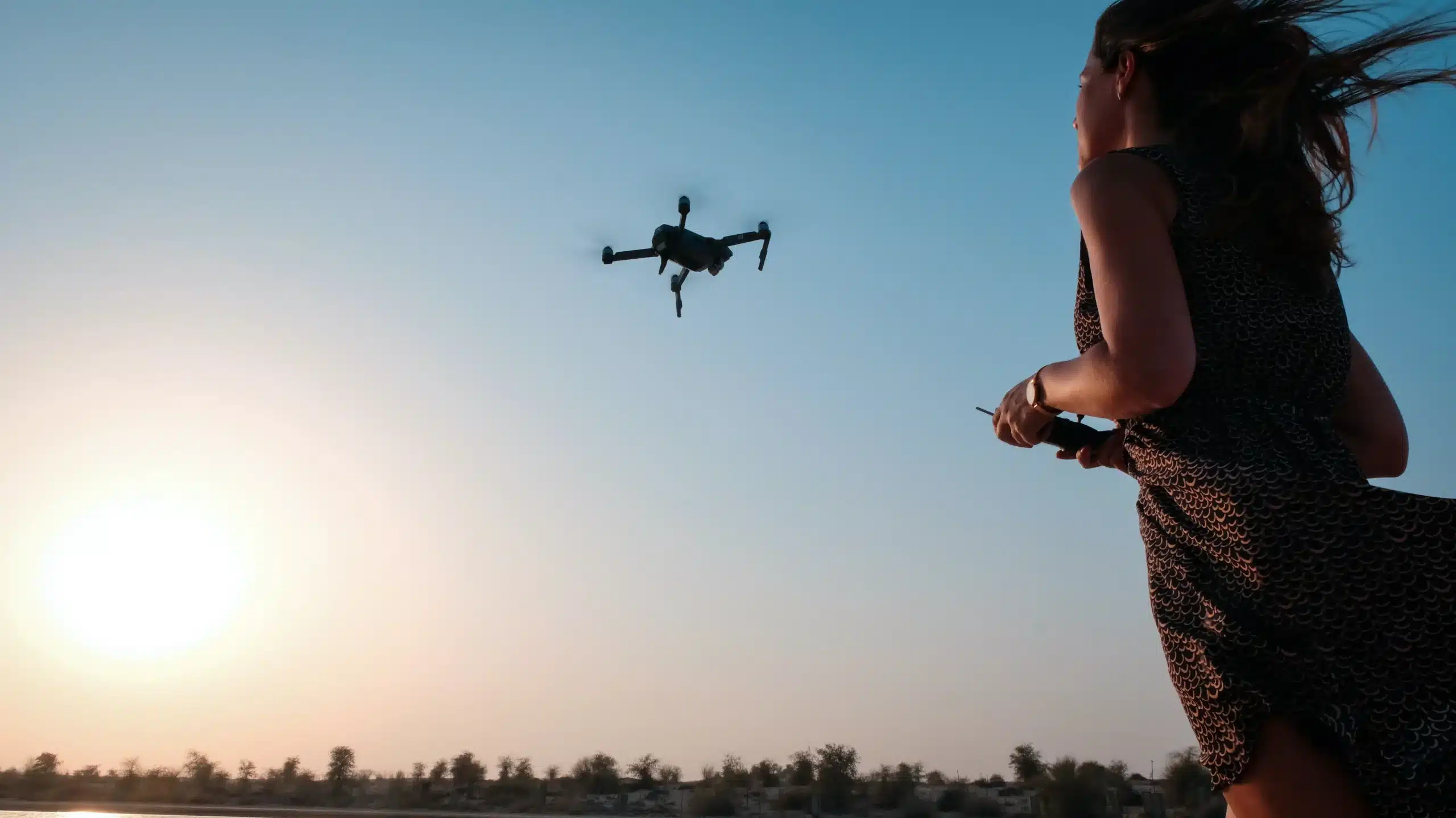 Eine Frau in einem gemusterten Kleid bedient eine fliegende Drohne während des Sonnenuntergangs, mit einem klaren blauen Himmel über der Segelyacht im Hintergrund.