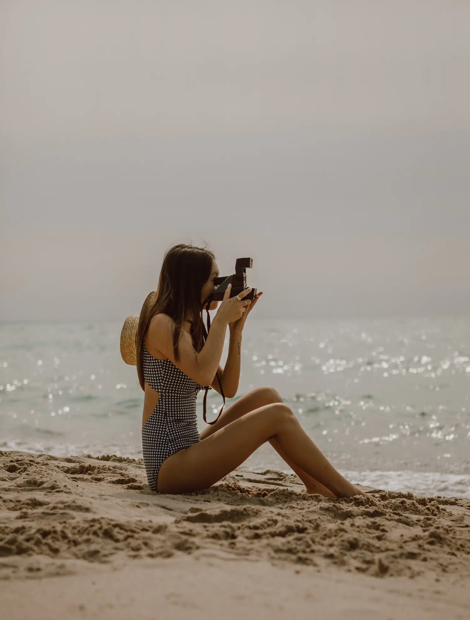Eine Frau in einem schwarz-weißen Badeanzug sitzt an einem Sandstrand und fotografiert während ihrer Segelreise mit ihrer Kamera das Meer. Sie trägt einen Strohhut und eine Brille, mit