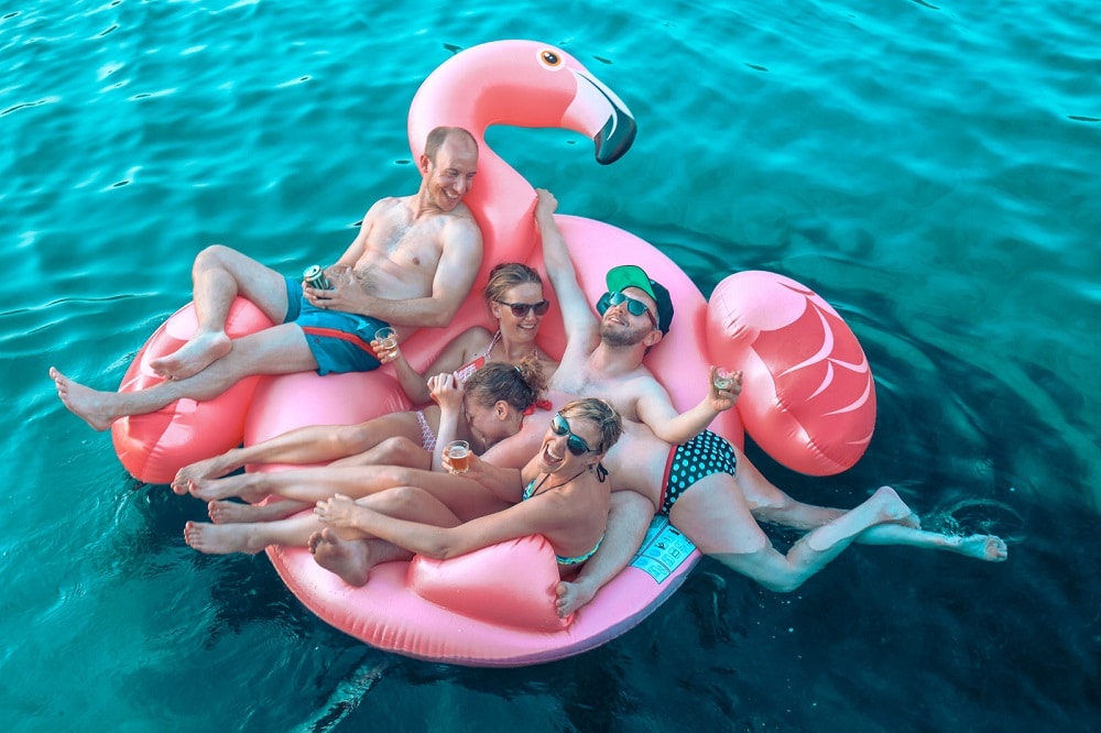 Vier Erwachsene entspannen sich und genießen Getränke auf einem großen rosa Flamingo-Schwimmreifen im klaren, blauen Wasser, lächeln und tanken Sonne beim Segeltörn.