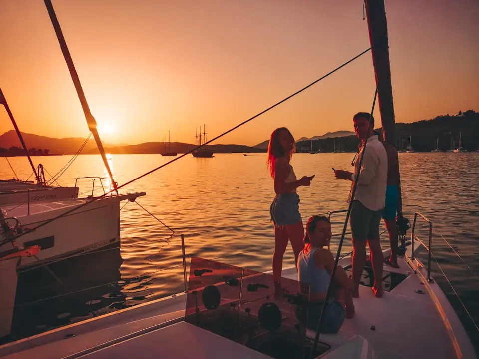 Drei Freunde genießen den Sonnenuntergang von einer Segelyacht aus. Einer sitzt, zwei stehen, halten Getränke in der Hand und unterhalten sich in einem ruhigen Hafen, der von warmem, goldenem Licht erhellt wird.