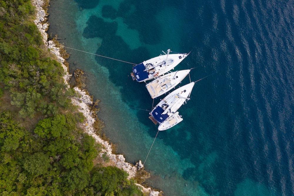 Luftaufnahme von zwei Katamaranen, die nebeneinander in der Nähe einer felsigen Küste mit üppiger grüner Vegetation vor Anker liegen und während eines Segeltörns auf dem klaren, türkisfarbenen Wasser treiben.