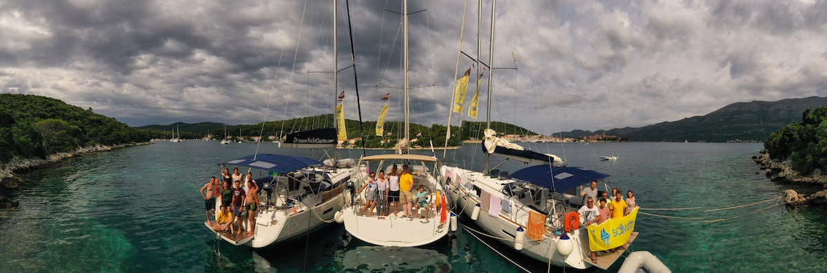 Panoramablick auf einen malerischen Ankerplatz mit drei zusammengebundenen Segelbooten, voll mit Menschen, die ein sonniges Segeln genießen, vor der Kulisse üppig grüner Hügel und eines bewölkten Himmels.