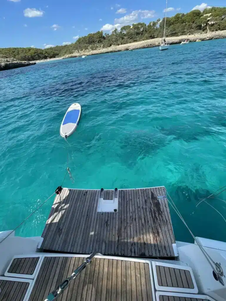 Der Blick vom Heck einer Segelyacht zeigt ein Holzdeck, das zum klaren, türkisfarbenen Wasser führt, mit einem kleinen weißen Boot, das in der Nähe schwimmt, umgeben von üppigem Grün und einer weiteren Yacht in der Ferne.