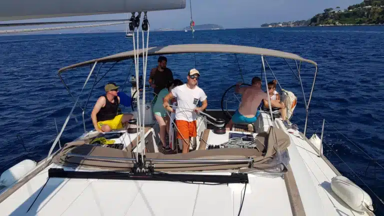 Eine Gruppe von Menschen genießt einen sonnigen Tag auf einem Segelboot während eines Segeltörns. Einige sitzen und stehen auf dem Deck, das Meer ist ruhig und im Hintergrund ist die Küste zu sehen.
