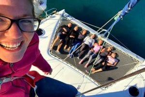 Eine Frau macht ein Selfie auf einer Segelyacht; im Vordergrund fällt ihr Lächeln auf, während im Hintergrund auf dem Deck eine Gruppe von Menschen entspannt und lächelnd liegt.