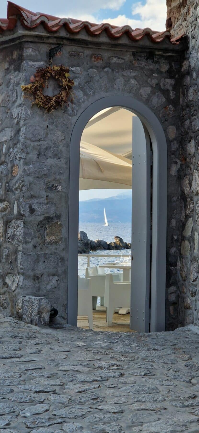 Blick durch einen gewölbten Steineingang, der zu einem Restaurant am Meer führt. In der Ferne ist auf dem Meer unter einem blauen Himmel eine weiße Segelyacht zu sehen.