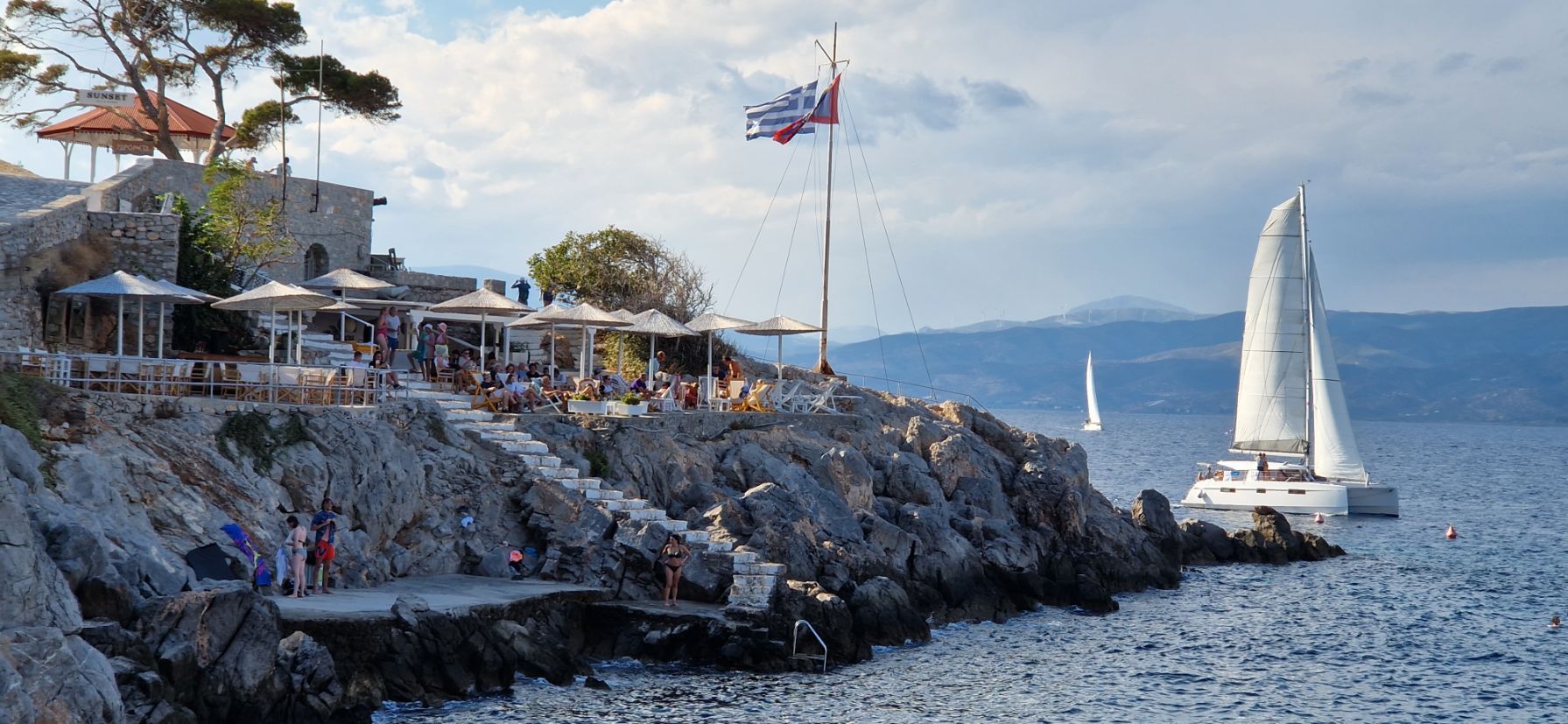 Ein Restaurant am Meer auf einem Felsvorsprung mit einer wehenden kroatischen Flagge. Menschen speisen im Freien, während in der Nähe ein Segelboot unter leicht bewölktem Himmel auf dem glitzernden Meer zu einem Segelurlaub aufbricht.