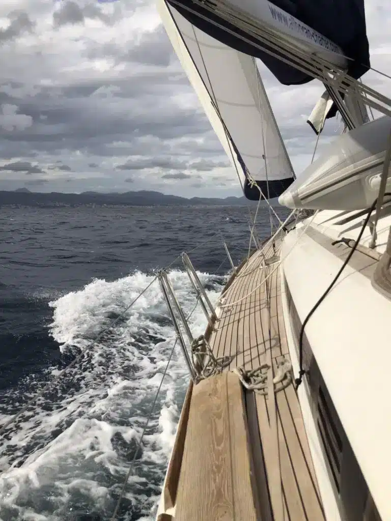 Ein Segelboot kreuzt auf offenem Wasser, sein Deck ist mit weißen Segeln unter einem bewölkten Himmel sichtbar, in der Ferne sind bergige Küstenlinien schwach zu erkennen. Genießen Sie einen Segeltörn