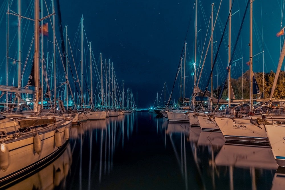 Eine ruhige Nachtszene in einem Yachthafen. Segelyachten liegen aufgereiht auf ruhigem Wasser unter einem Sternenhimmel und werden vom sanften Licht des Docks beleuchtet.