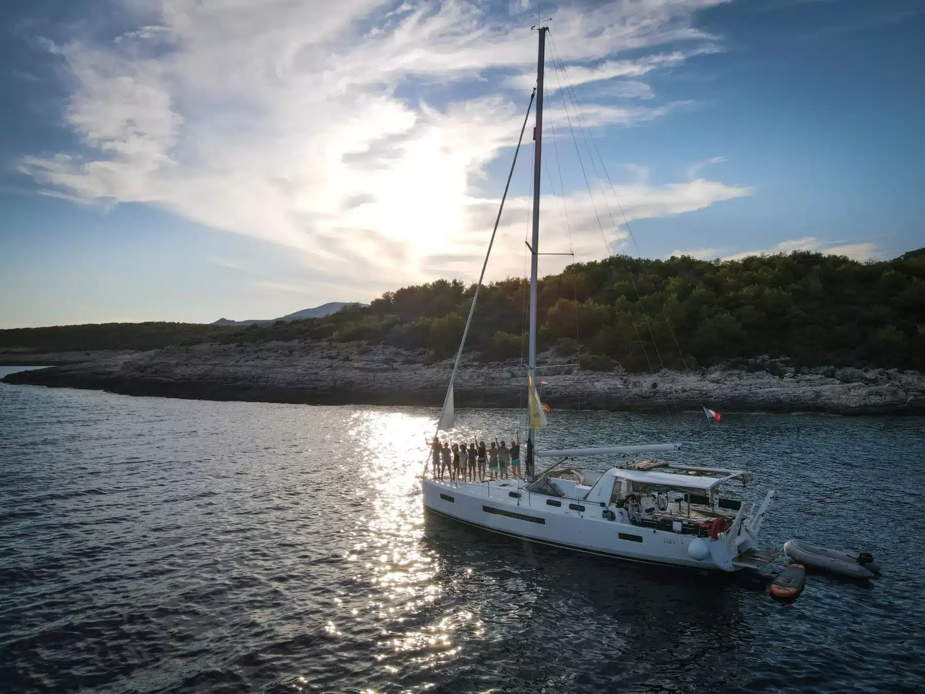 Ein Segelboot mit mehreren Personen an Bord liegt in der Nähe einer felsigen Küste vor Anker, unter einem sanften Sonnenuntergangshimmel. Hinter ihnen zieht ein kleines Schlauchboot auf ihrer Segelreise her.