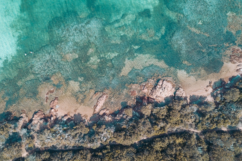 Luftaufnahme einer Küste, die das klare blaue Wasser des Meeres zeigt, das in flache Riffe und Sandstrände übergeht, wobei während eines Segeltörns subtile Texturen und Farben unter Wasser sichtbar sind.