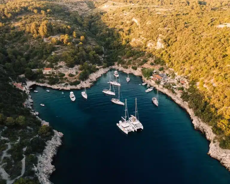 Luftaufnahme einer ruhigen Bucht mit türkisfarbenem Wasser, umgeben von einem üppigen grünen Wald, mit Yachten und Booten, die während ihres Segelurlaubs in der Nähe der zerklüfteten Küste vor Anker liegen.