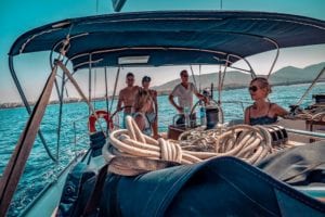 Eine Gruppe von Freunden genießt einen sonnigen Tag auf einer Segelyacht mit klarem blauen Himmel und Bergen im Hintergrund. Ein Mann steuert, während die anderen sich entspannen und unterhalten.