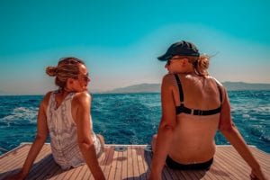 Zwei Frauen sitzen während eines Segeltörns auf dem Achterdeck eines Bootes und blicken unter einem klaren blauen Himmel auf das Meer. Eine Frau trägt ein weißes Oberteil und die andere einen schwarzen Bikini