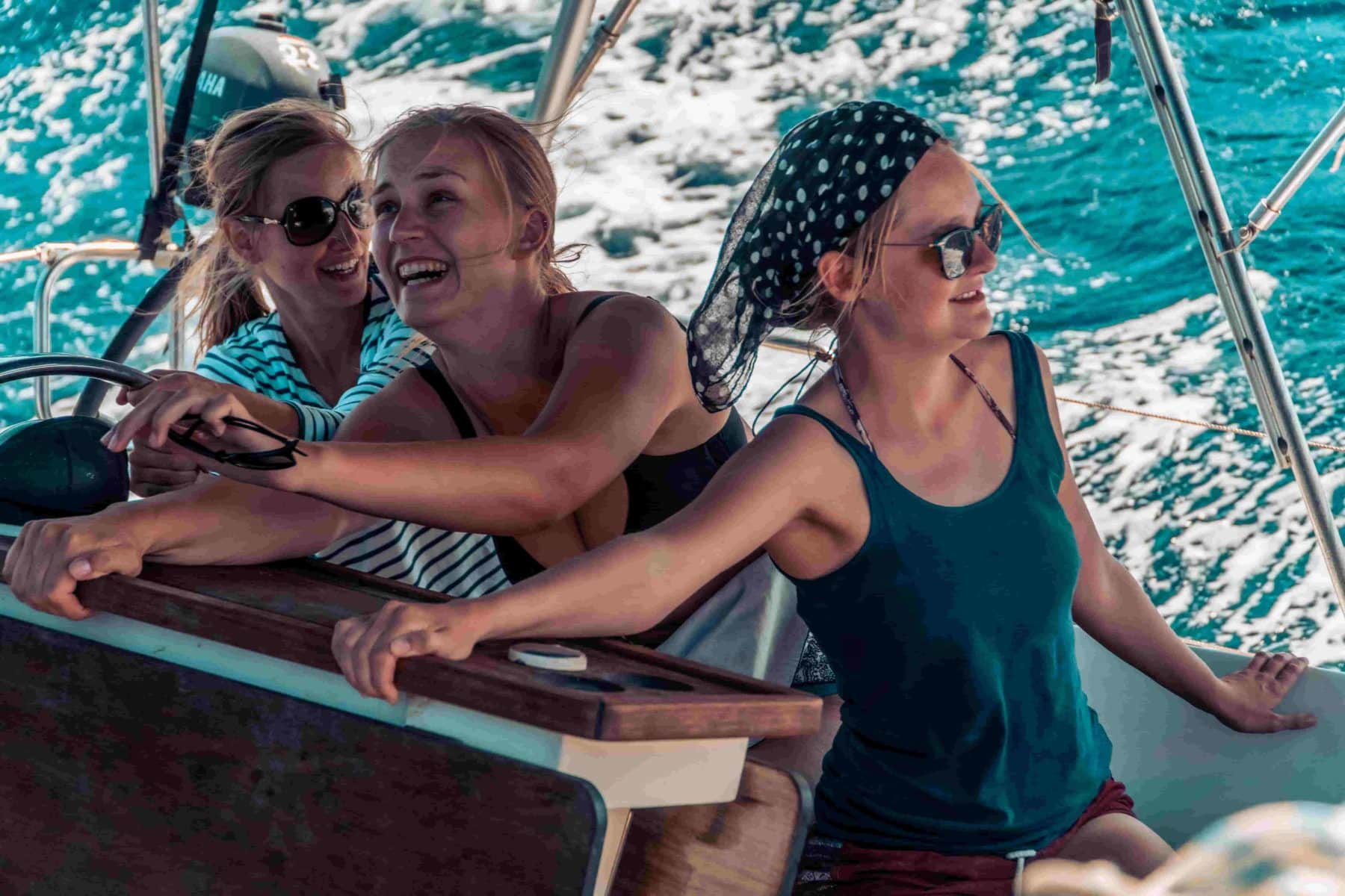Drei Frauen lachen und genießen einen sonnigen Tag beim Segeln, zwei stehen am Steuer und eine steht daneben, alle sind leger gekleidet und tragen Sonnenbrillen.