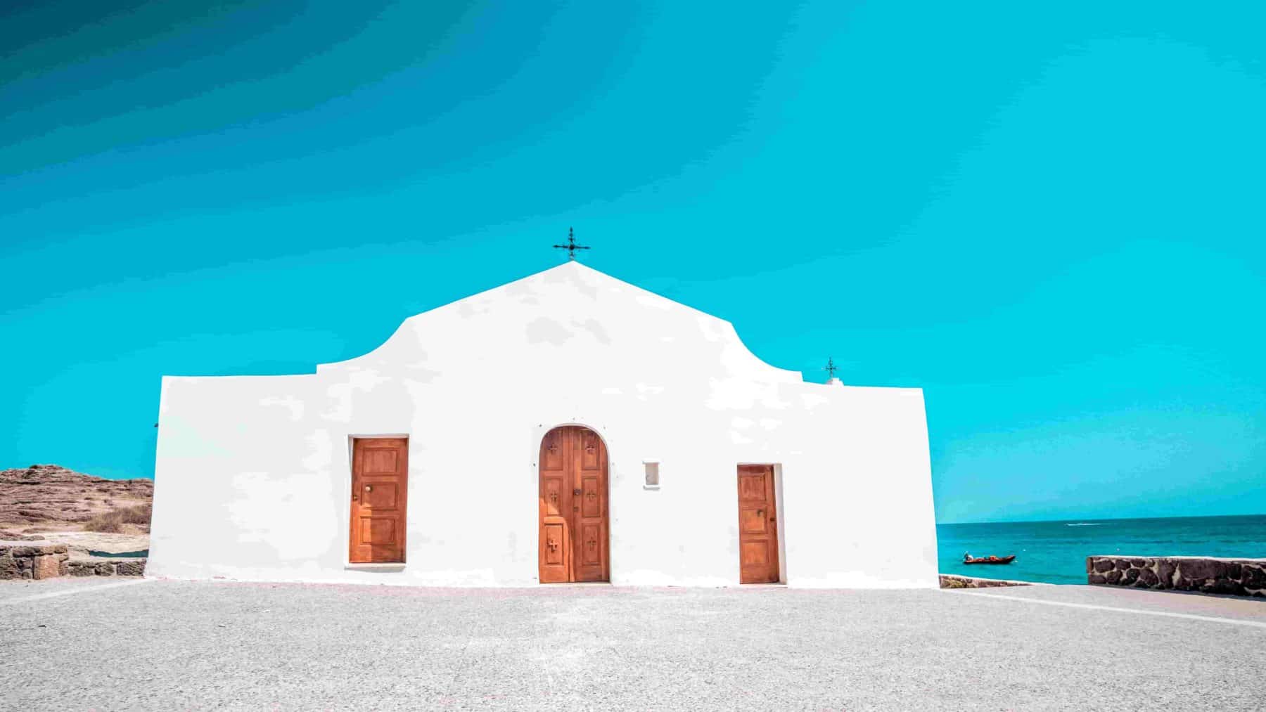 Eine kleine weiße Kapelle mit einer einfachen, geschwungenen Fassade und Holztüren unter einem klaren blauen Himmel, gelegen am Meer, mit einer zerklüfteten Küstenlinie und mehreren Segelyachten im Hintergrund.