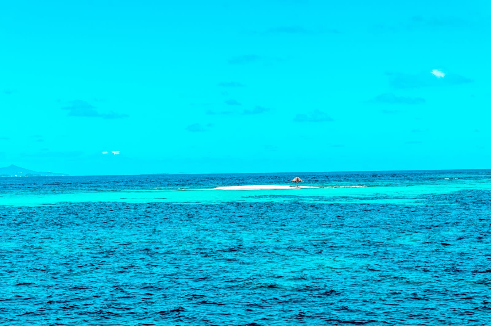 Ein leuchtend türkisfarbener Ozean mit einer kleinen, entfernten Segelyacht auf einer Sandbank unter einem klaren blauen Himmel.