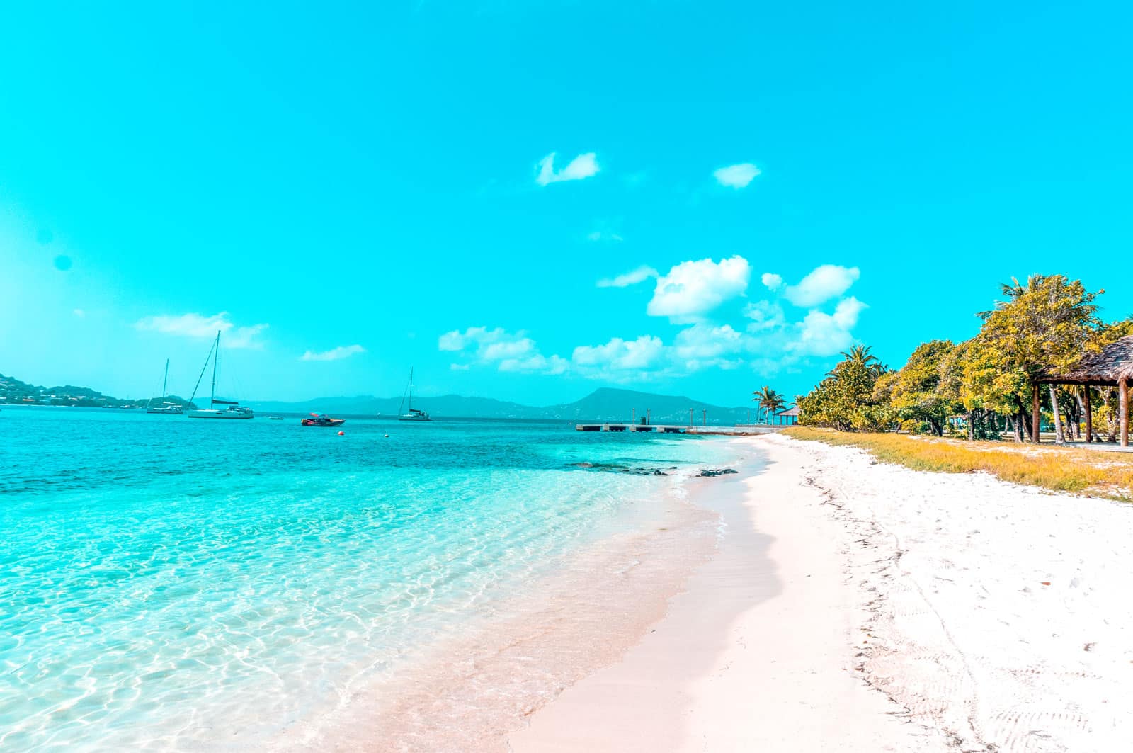 Eine lebendige tropische Strandszene mit leuchtend türkisfarbenem Meer, weißem Sandstrand, verstreuten Segelyachten und üppigem grünem Laub unter einem klaren blauen Himmel mit flauschigen Wolken.