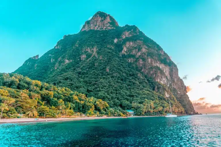 Ein majestätischer Berg überblickt einen tropischen Strand mit üppigem Grün und einem ruhigen blauen Meer bei Sonnenuntergang. Ein einsames Segelboot schwimmt in Ufernähe unter einem Himmel in sanften Pastellfarben.