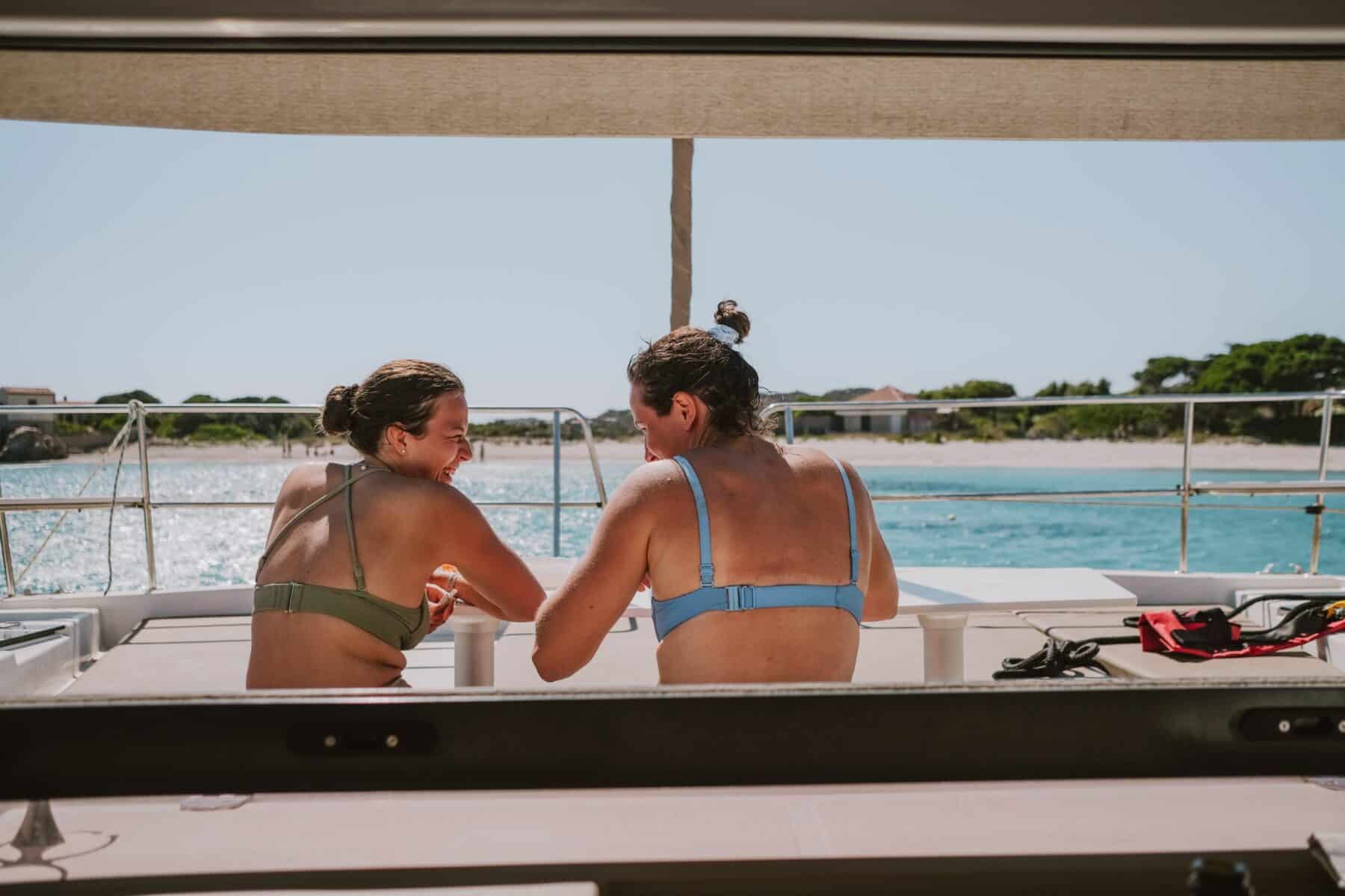 Zwei Personen in Badeanzügen sitzen und unterhalten sich auf dem Heck eines Segelboots und blicken auf ein ruhiges blaues Meer unter einem klaren Himmel.