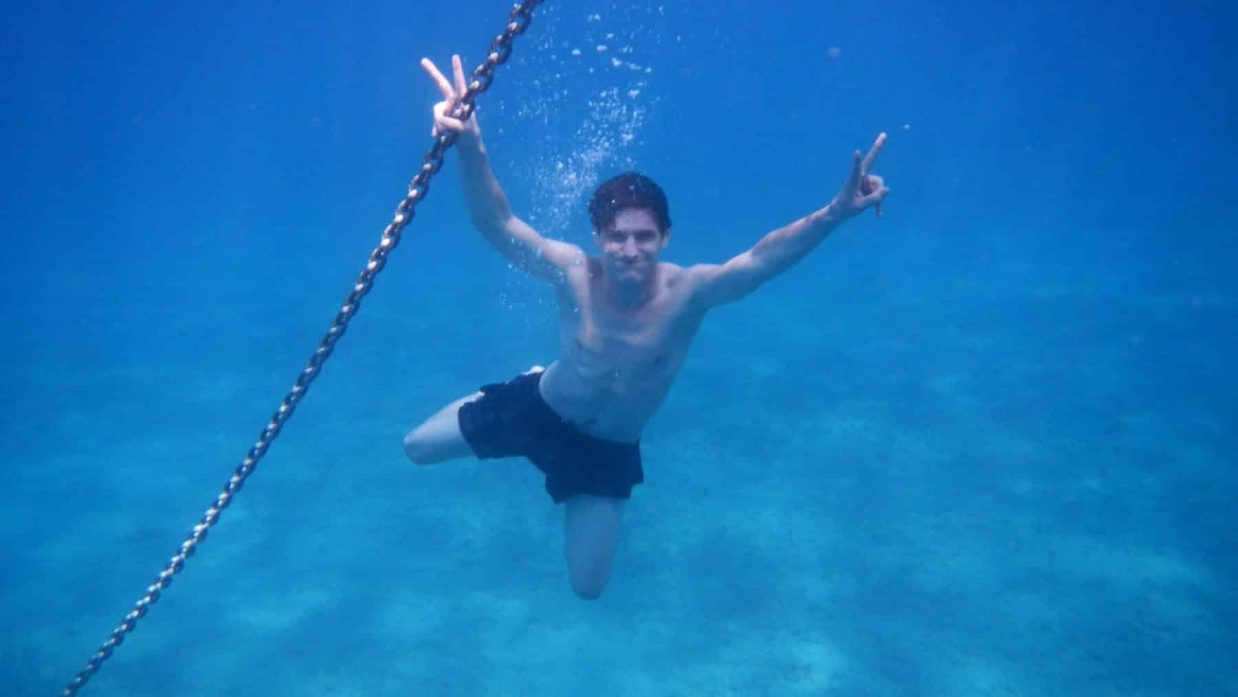 Ein Mann unter Wasser macht mit beiden Händen Peace-Zeichen und schwimmt neben einer vertikalen Kette einer Segelyacht im klaren, blauen Wasser.