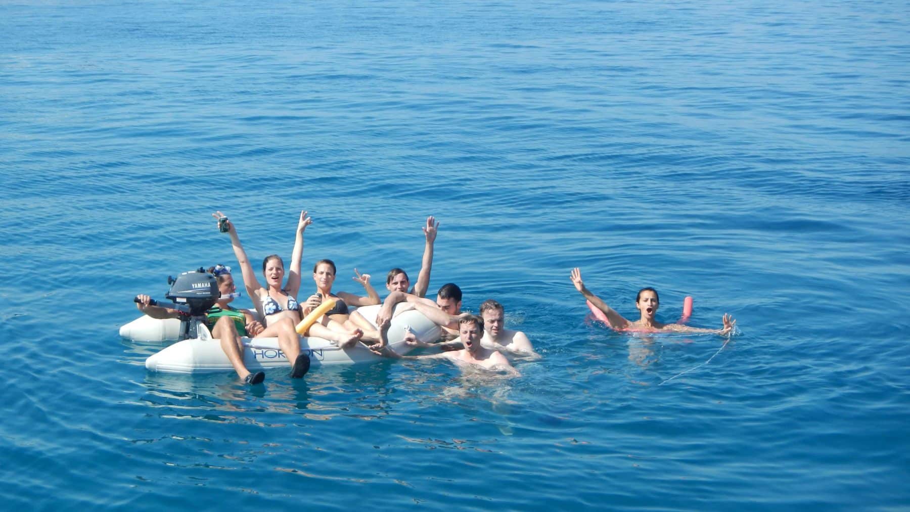 Eine Gruppe von sieben Menschen treibt fröhlich auf einer Segelyacht im klaren, blauen Ozean, einige winken und zeigen Peace-Zeichen und genießen einen sonnigen Tag.