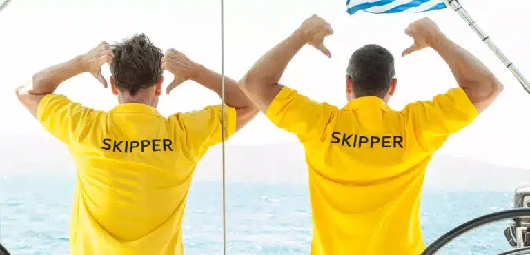 Zwei Männer in gelben "Skipper"-Hemden stehen Rücken an Rücken auf einem Boot, jeder gestikuliert mit dem Daumen nach oben, während er sich am Bootsrahmen festhält, mit dem Meer im Hintergrund während eines Seg