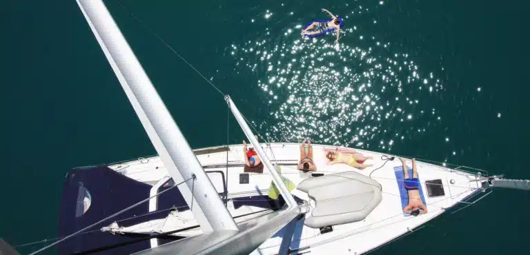 Luftaufnahme einer Segelyacht, die auf klarem, blauem Wasser schwimmt, mit vier Personen, die auf dem Deck ein Sonnenbad nehmen, und einer Person, die in der Nähe schwimmt.