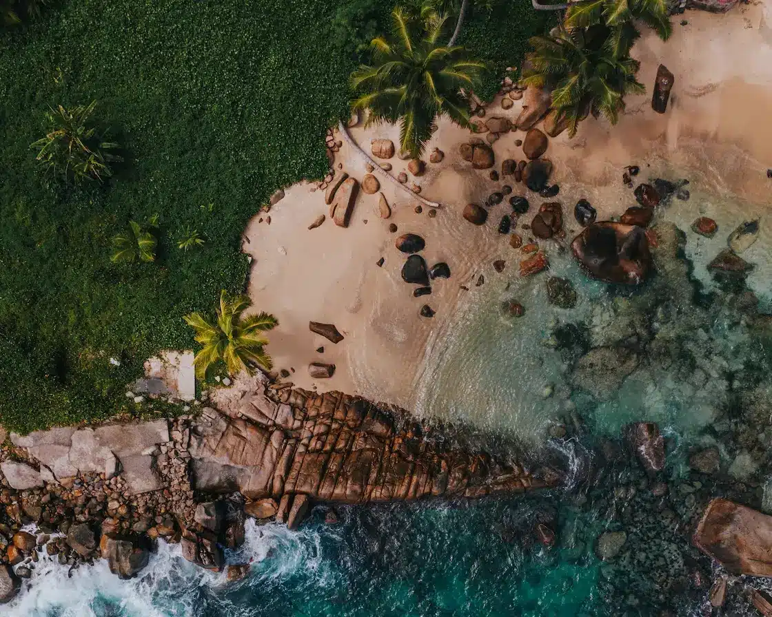 Luftaufnahme eines tropischen Strandes mit klarem türkisfarbenem Wasser, weißem Sand, verstreuten Felsen und üppig grünen Palmen, perfekt für einen Segeltörn.