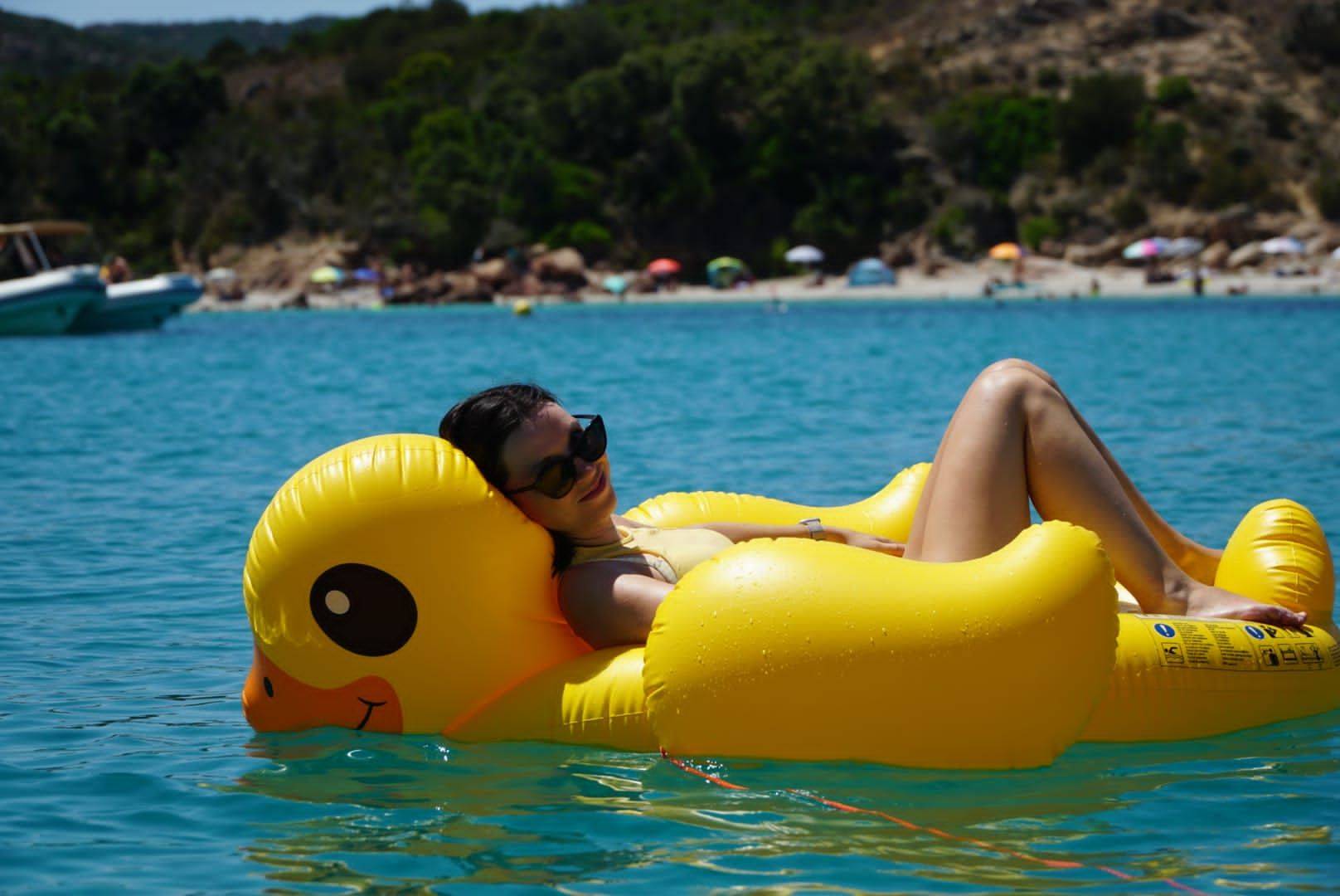 Eine Person, die auf einem großen, gelben, aufblasbaren Schwimmkörper in Entenform im klaren, blauen Wasser liegt. Im Hintergrund sind ein Sandstrand und Segelboote zu sehen.