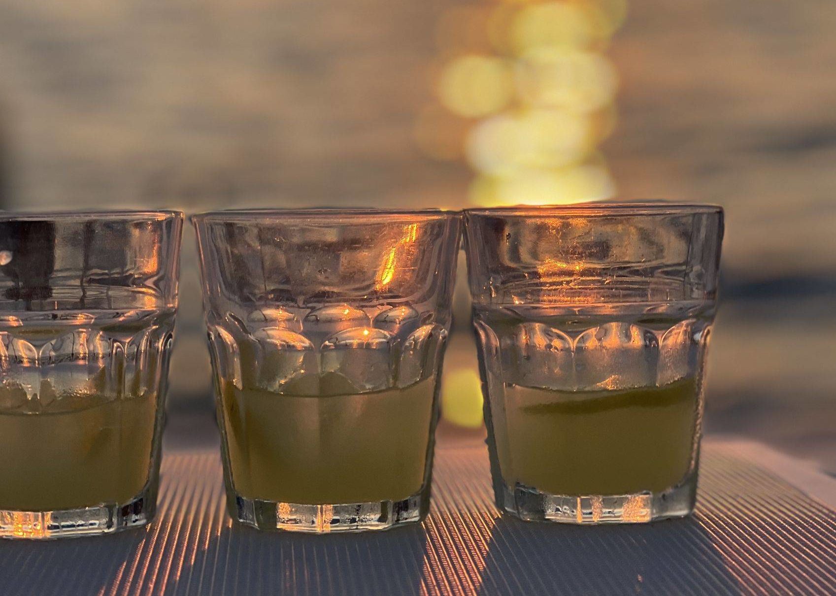 Drei mit einer goldenen Flüssigkeit gefüllte Gläser stehen auf einem Tisch an Bord einer Segelyacht, beleuchtet vom warmen Licht des Sonnenuntergangs, das sich im Wasser im Hintergrund spiegelt.