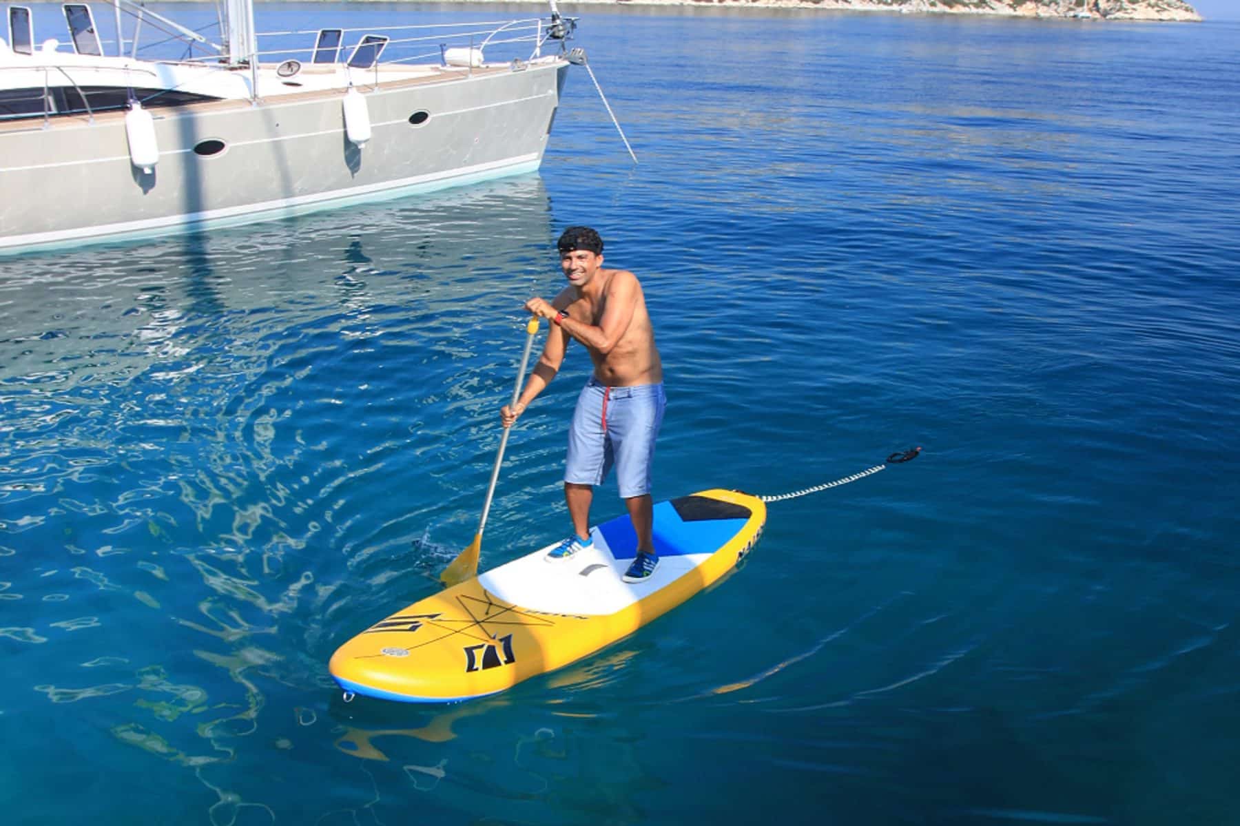 Ein Mann steht auf einem gelben Paddleboard im klaren blauen Wasser neben einer Segelyacht. Er trägt eine Mütze, eine Sonnenbrille, blaue Shorts und eine graue Schwimmweste und lächelt, während er paddelt.