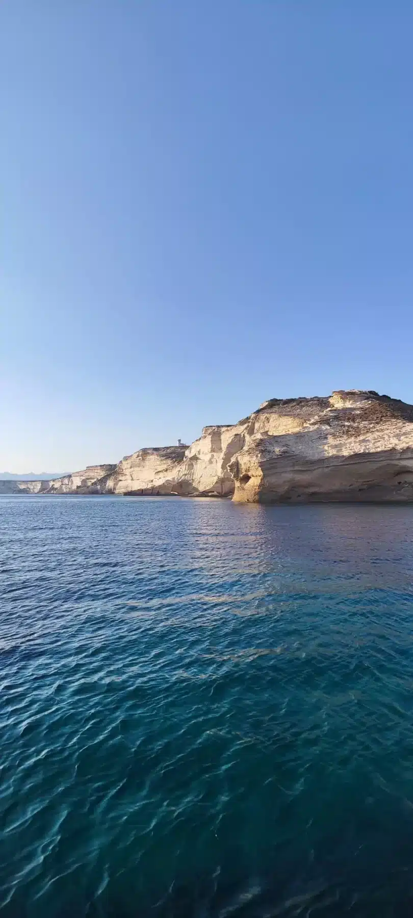 Eine ruhige Meereslandschaft mit klarem, blauem Wasser unter einem strahlenden Himmel und schroffen, geschichteten Sandsteinklippen am Meer während eines Segeltörns.